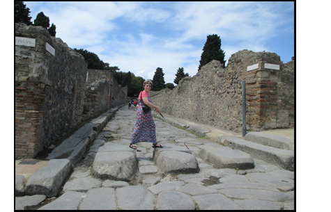 Photo shows Dona crossing a street in Pompeii, which has a row of 3 stepping stones, each about a foot high, 2 feet deep and 3 feet wide.  She is using a white cane and smiling at us.